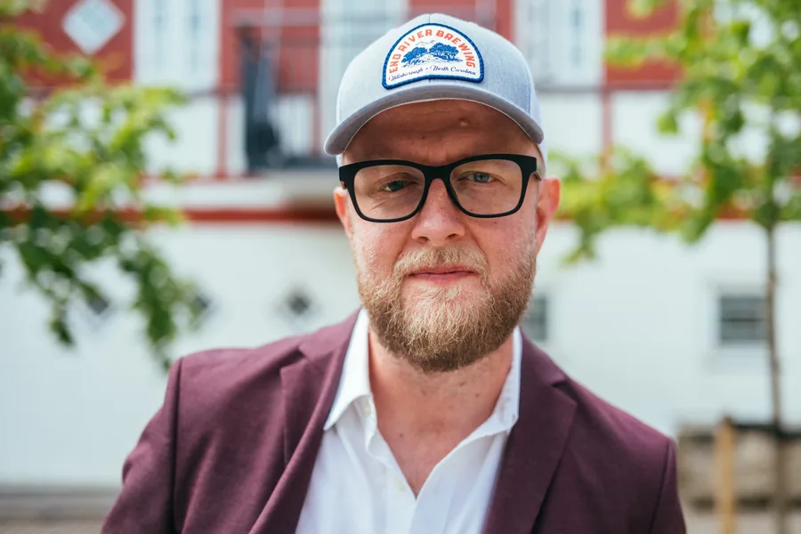 Portræt af Mikkel Elbech i kasket, briller og blommefarvet jakke