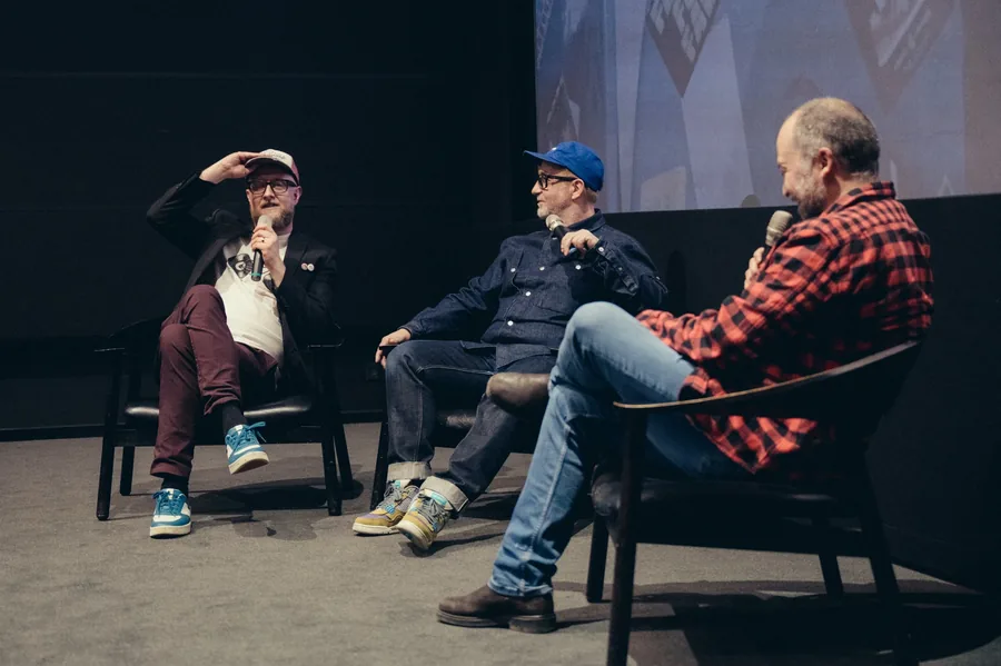 Tre mænd i stole på en scene med mikrofoner foran et biograflærred